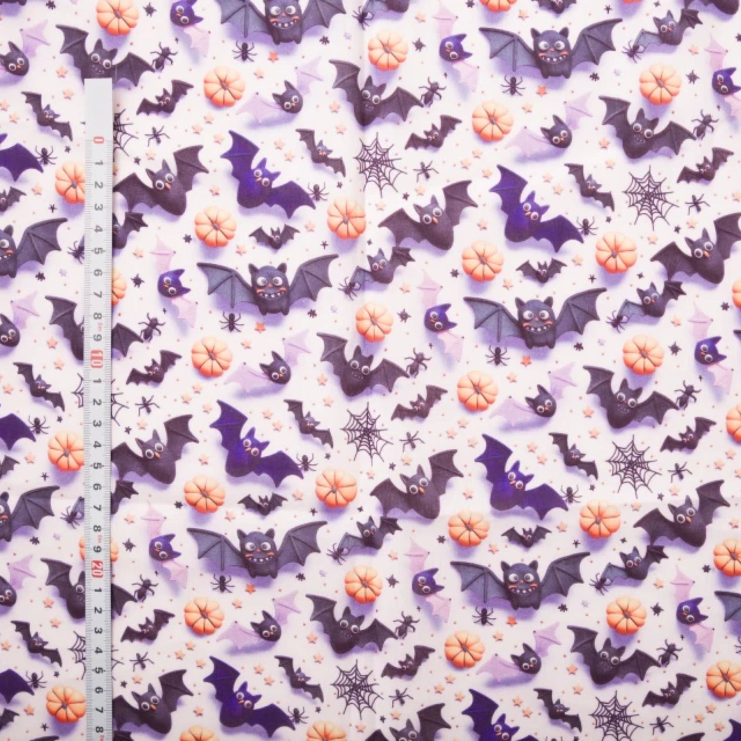 Halloween-themed fabric with a repeating pattern of purple bats, orange pumpkins, black spiderwebs, and small black stars on a white background, shown with a metal ruler on the left for scale.
