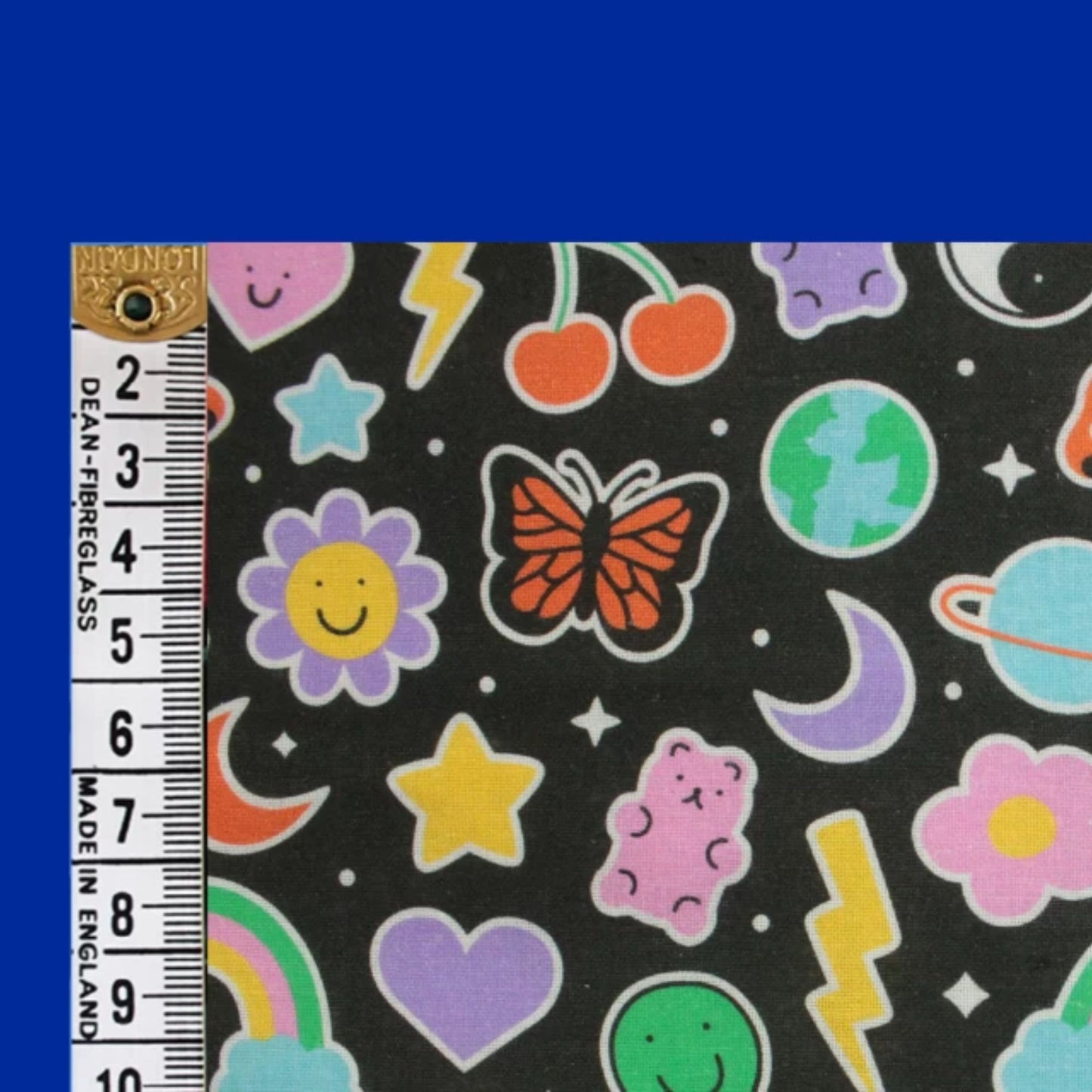 A close-up of a black fabric with a colorful cartoon space and nature print, shown next to a ruler for scale. The print includes a butterfly, flowers, stars, planets, and lightning bolts in pink, purple, yellow, green, orange, and blue.
