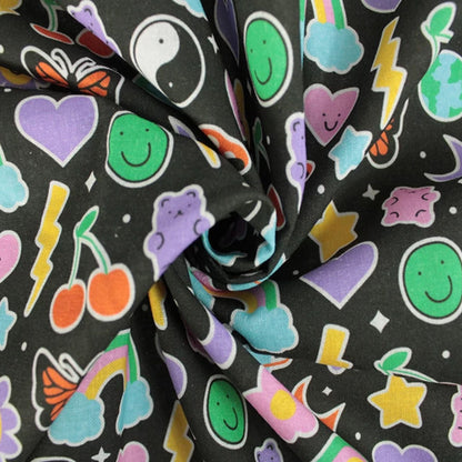 A close-up of a black fabric with a colorful cartoon pattern featuring smiley faces, hearts, stars, cherries, lightning bolts, rainbows, and other playful motifs.