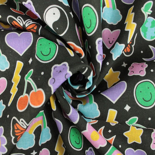 A close-up of a black fabric with a colorful cartoon pattern featuring smiley faces, hearts, stars, cherries, lightning bolts, rainbows, and other playful motifs.