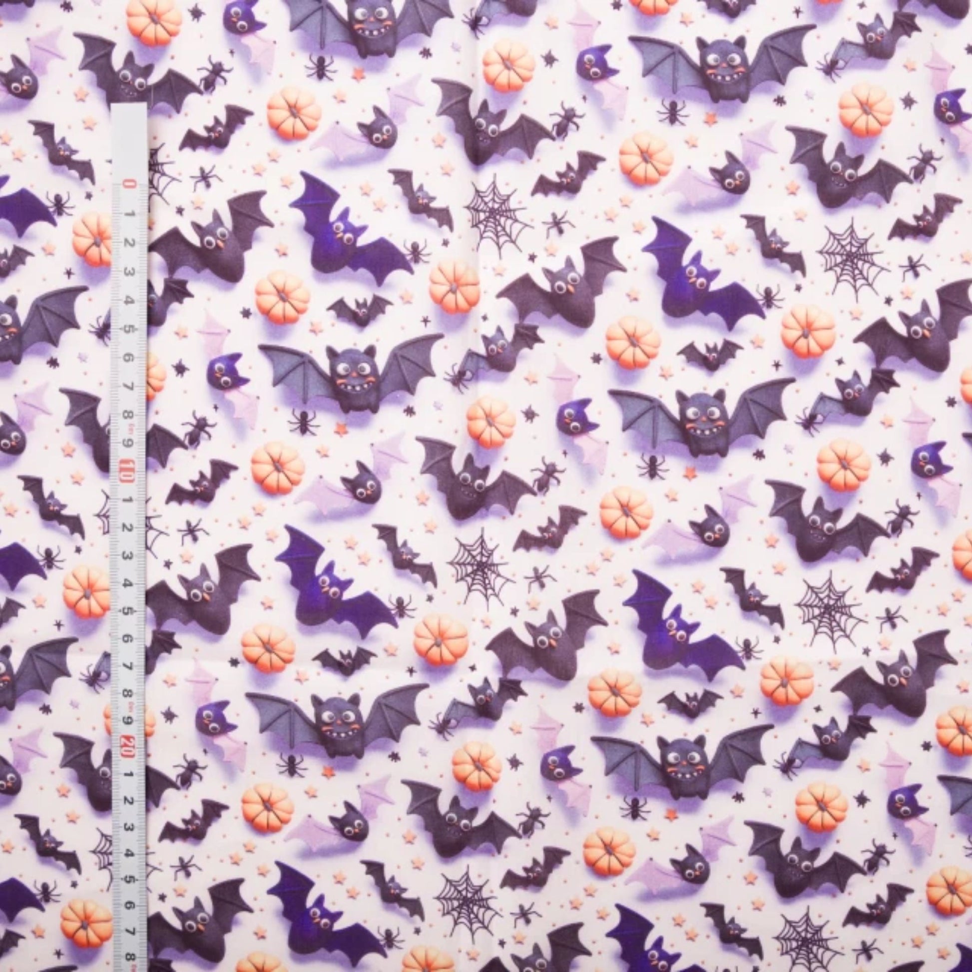 Halloween-themed fabric with a repeating pattern of purple bats, orange pumpkins, black spiderwebs, and small black stars on a white background, shown with a metal ruler on the left for scale.