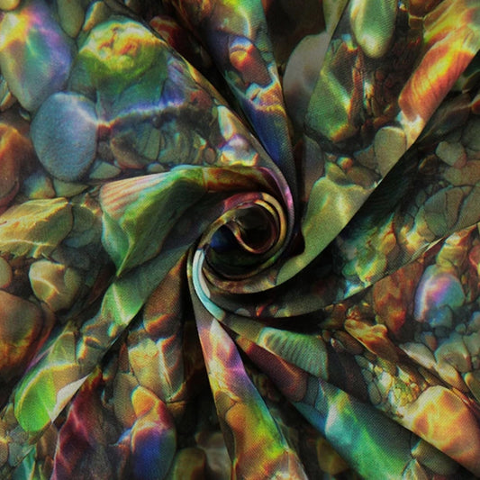 A close-up of iridescent fabric with a swirling, spiral pattern of pebbles. The material has a highly reflective, holographic finish showing shifting colors of green, purple, gold, blue, and orange over an abstract stone-like texture.
